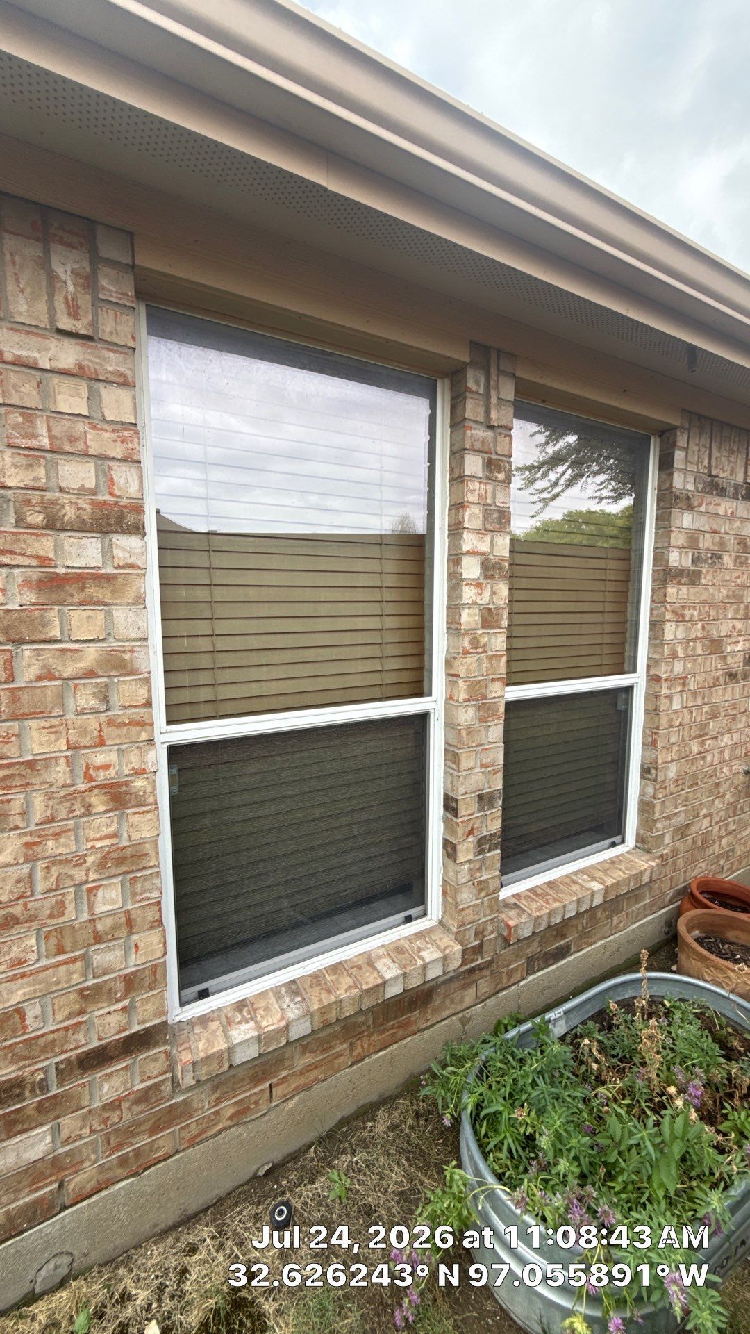 Brick house exterior with two windows, tan siding, and a small garden bed below.