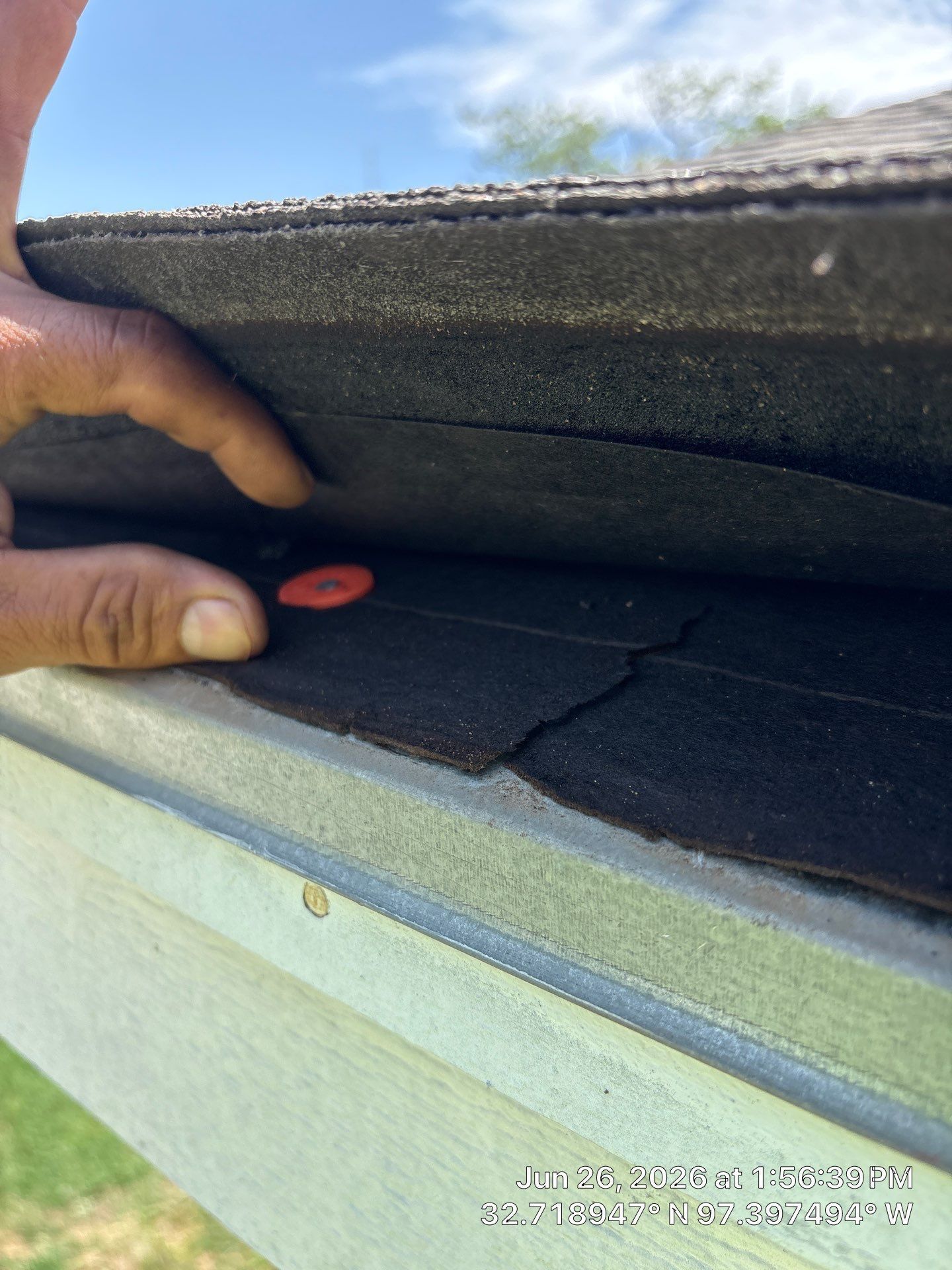 Hand holding a red tool to inspect dark roof shingles along a roof edge