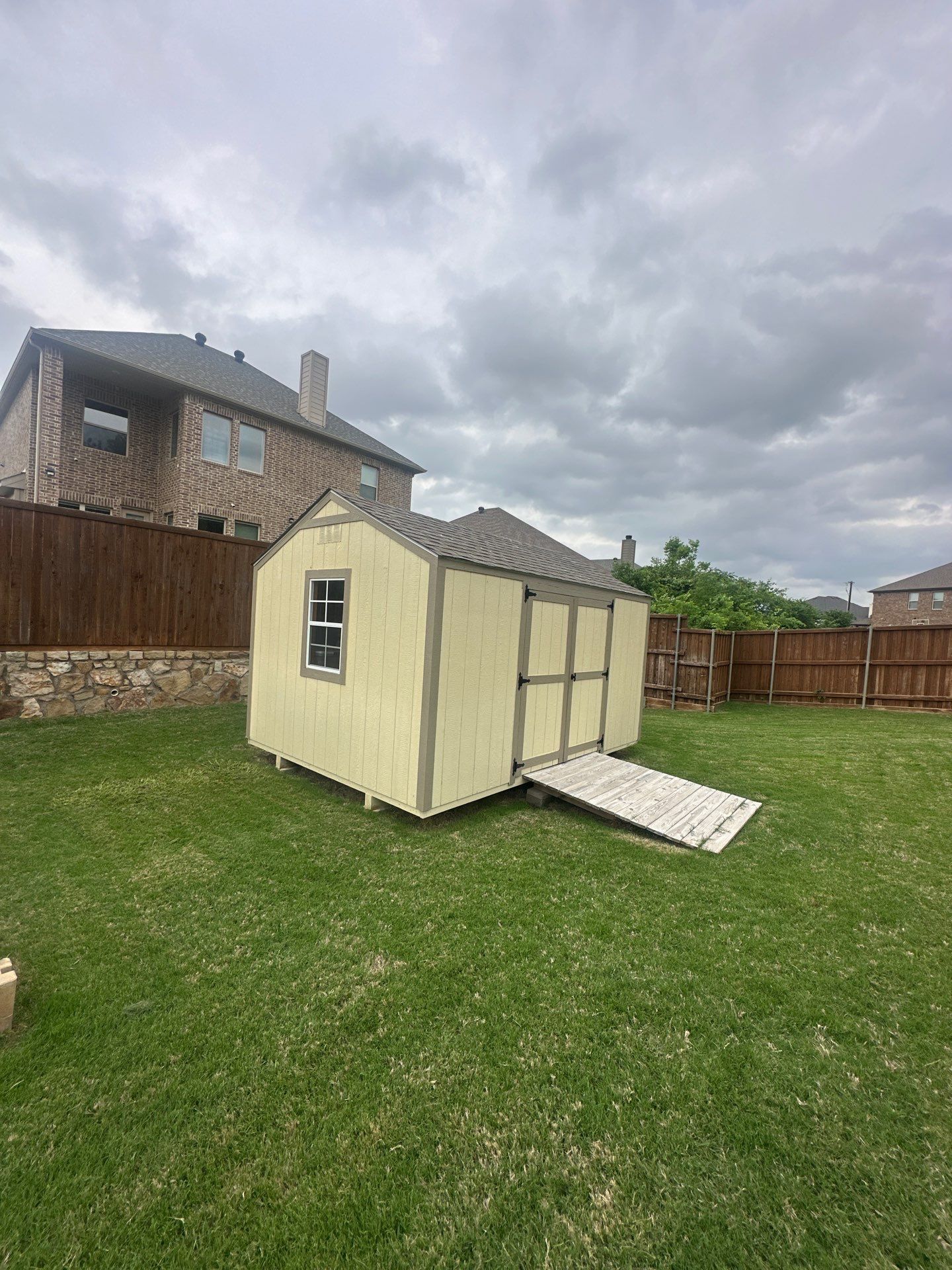 Small beige shed with a ramp in a grassy backyard under cloudy skies