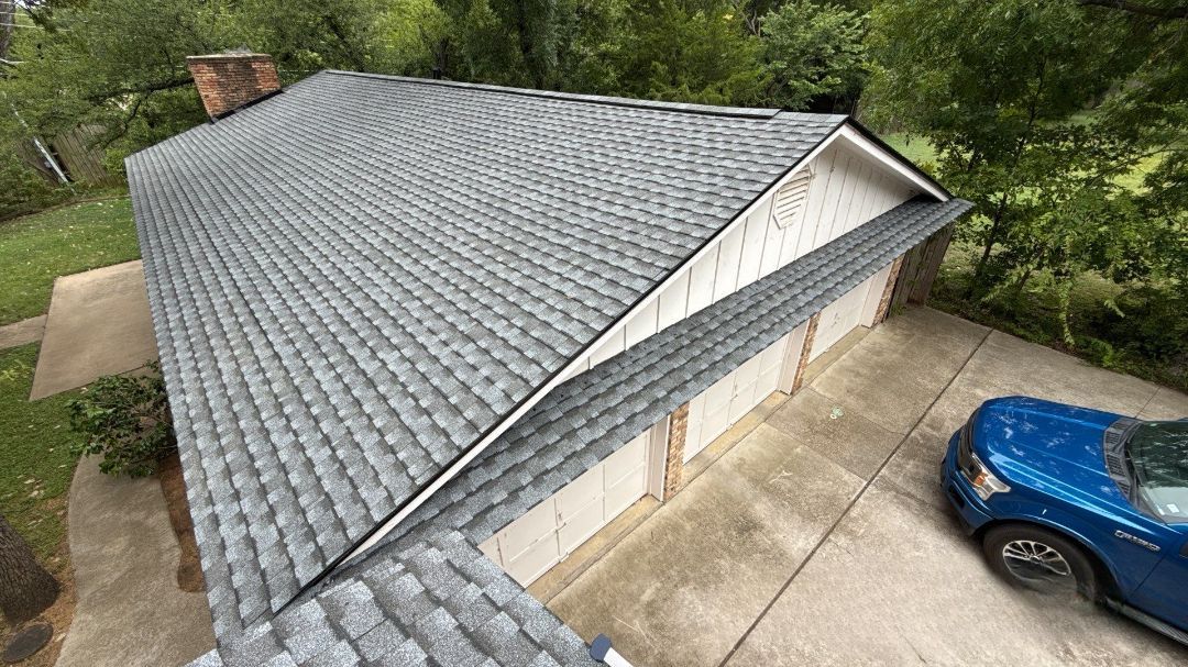 Aerial view of a gray-shingled house roof beside a driveway with a blue car and trees nearby