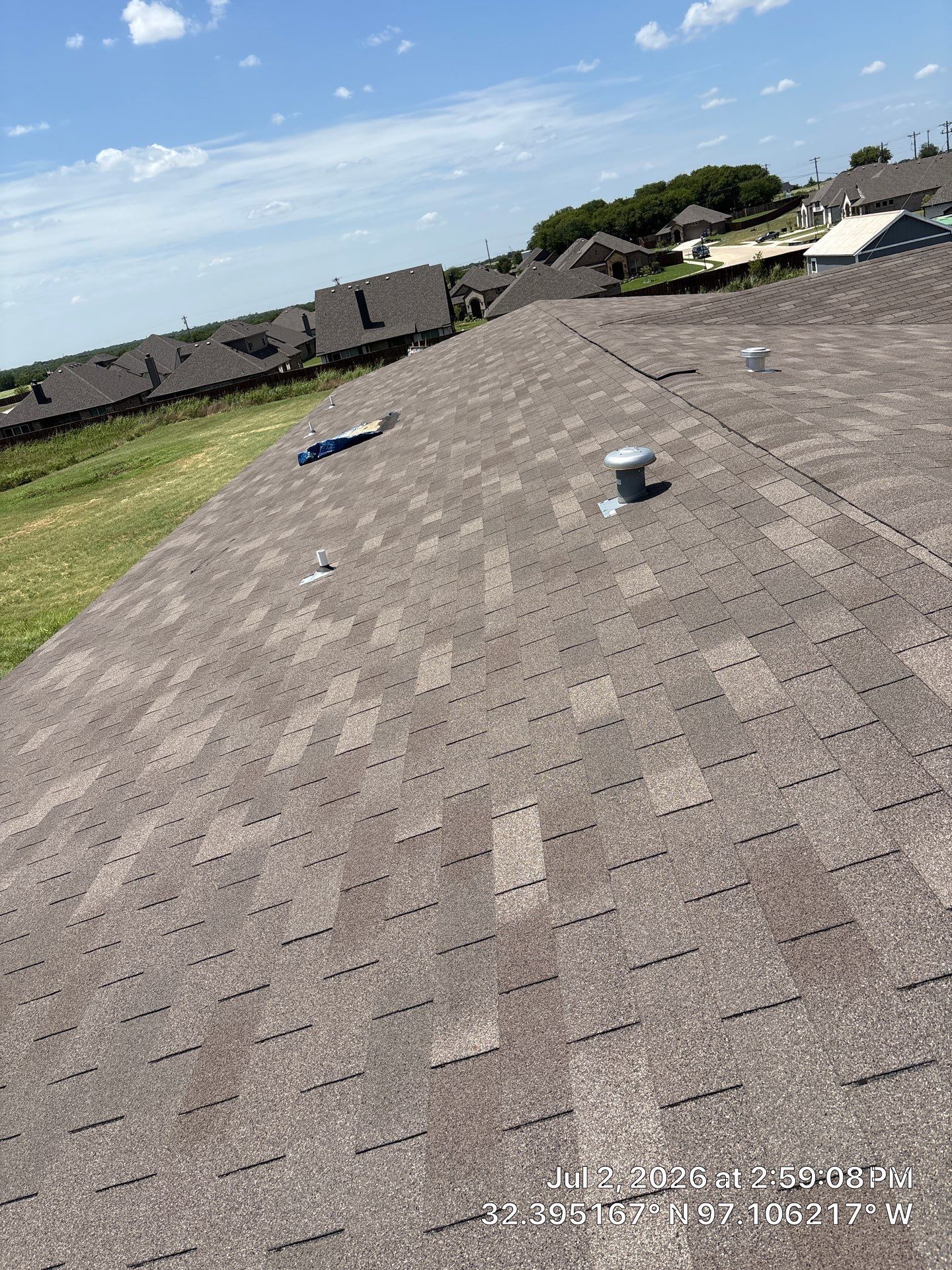 Rooftop with gray shingles and two vents under a blue sky, viewed from a slanted angle.