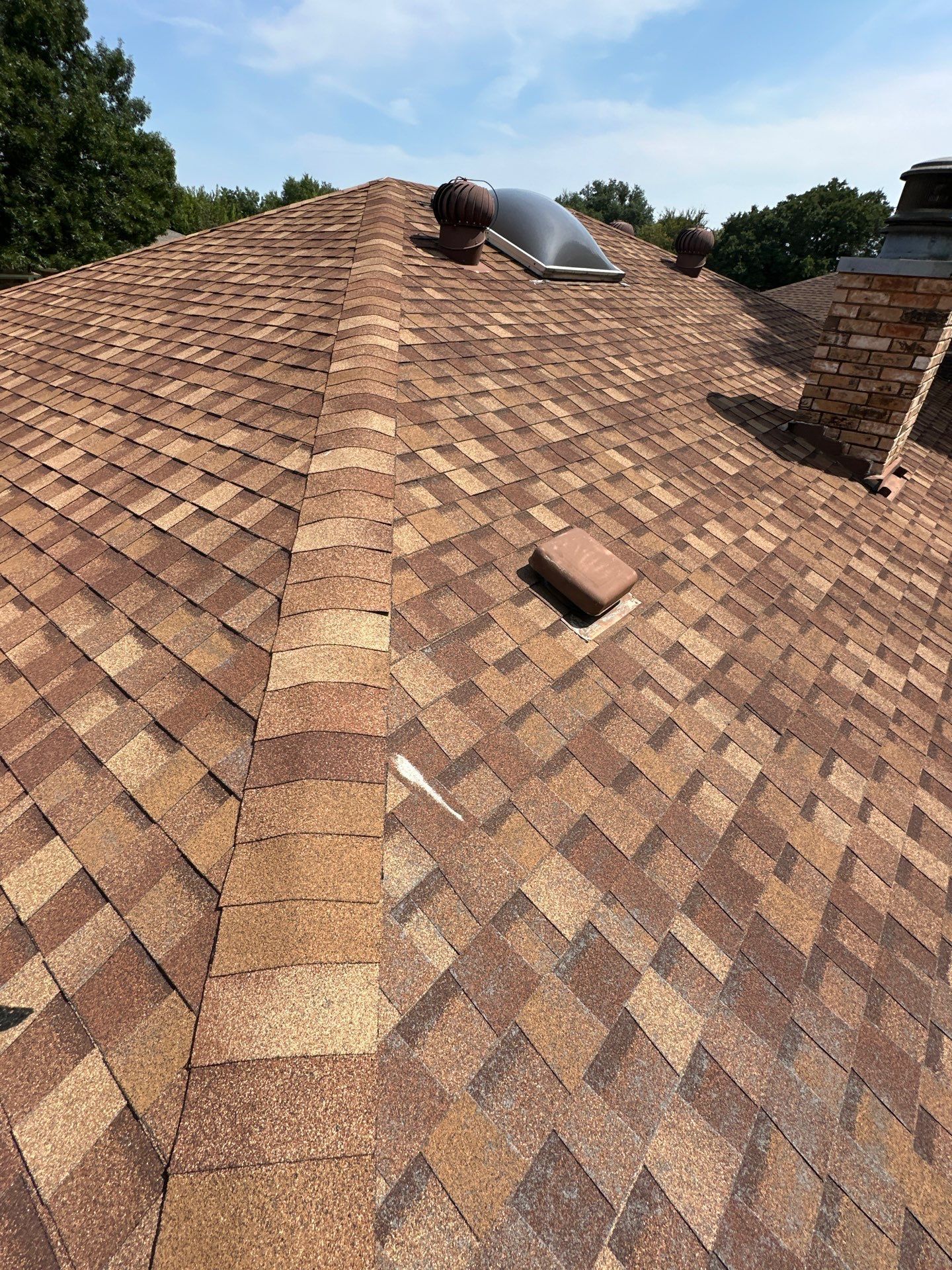 Roof with brown shingles and a vent pipe, viewed from above, with trees in the background.