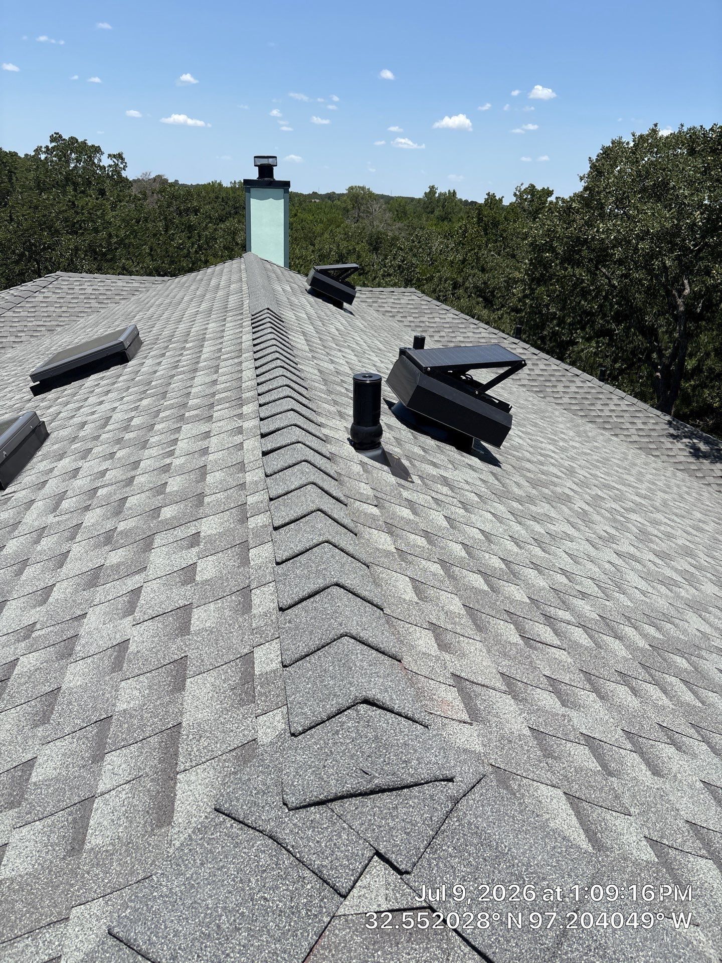 Roof with gray shingles, black vents, and a small chimney under a blue sky.