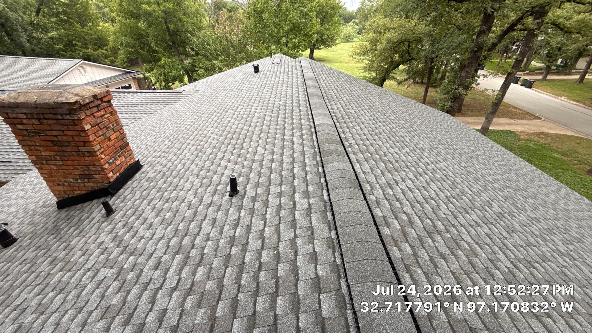 Roof covered in gray shingles with a brick chimney, viewed from above with trees in the background
