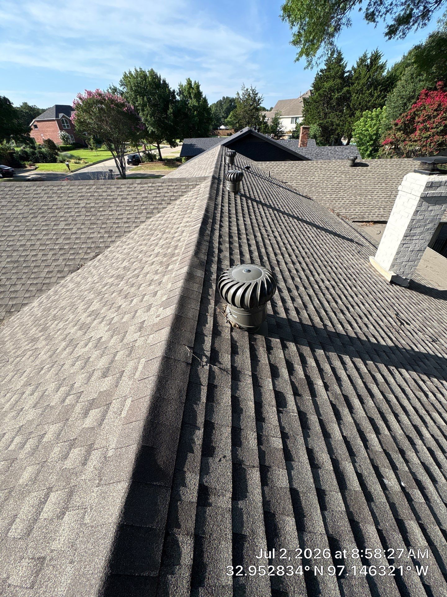 Roof with gray shingles, ridge vent, and chimney caps, viewed across a suburban neighborhood on a sunny day