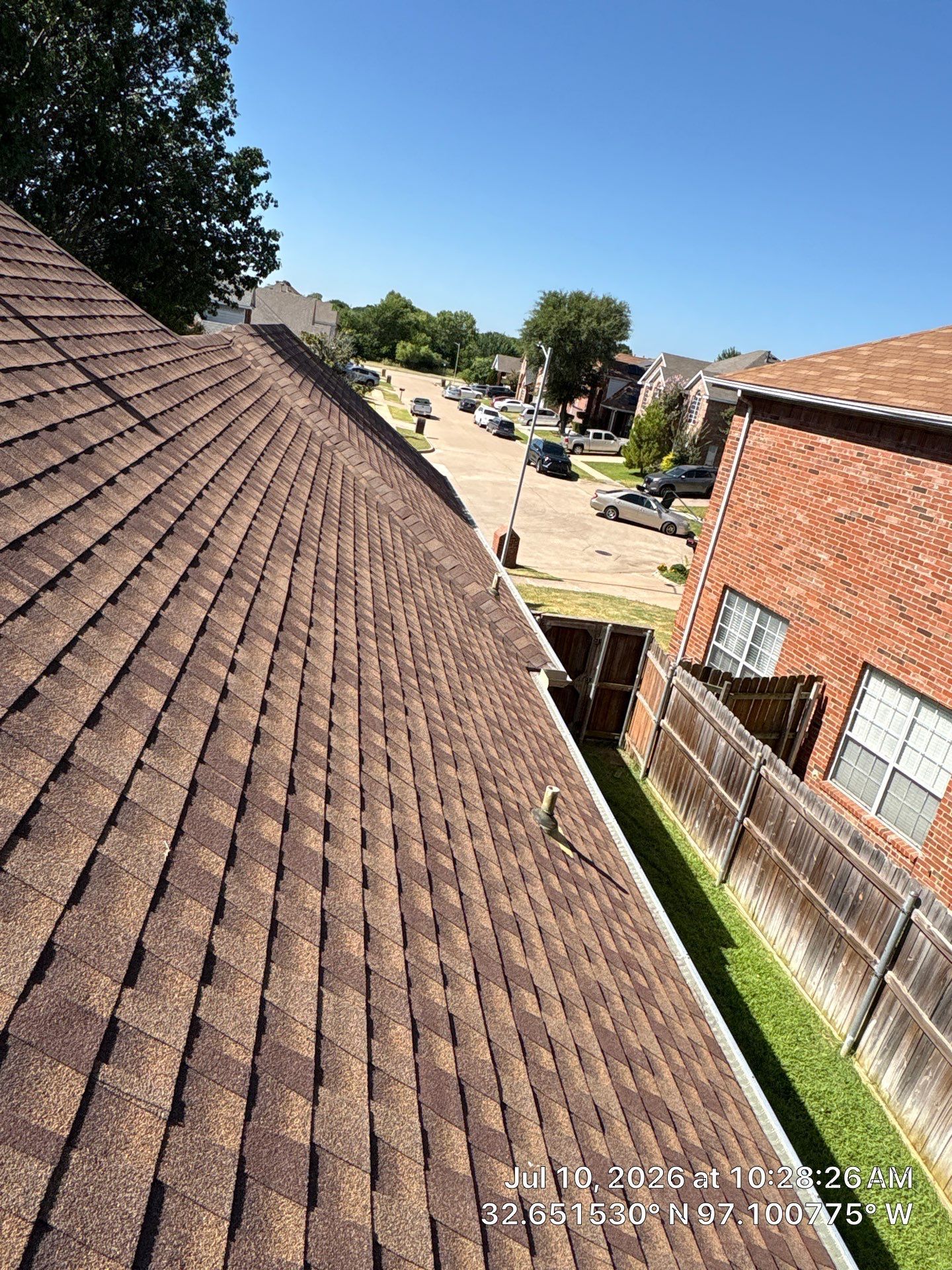 View from a shingled rooftop over a suburban street, with brick houses, a fenced yard, and bright blue sky