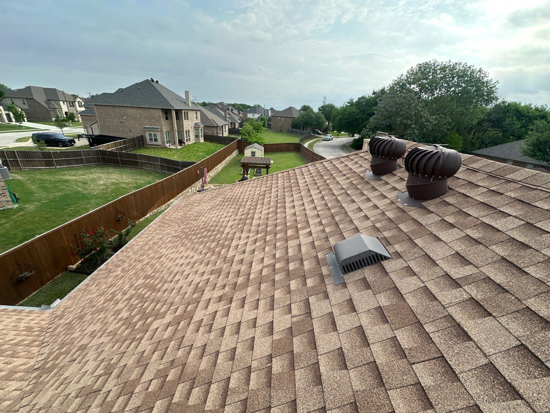 Rooftop view of a suburban neighborhood with brown shingles, vents, and backyards under a cloudy sky