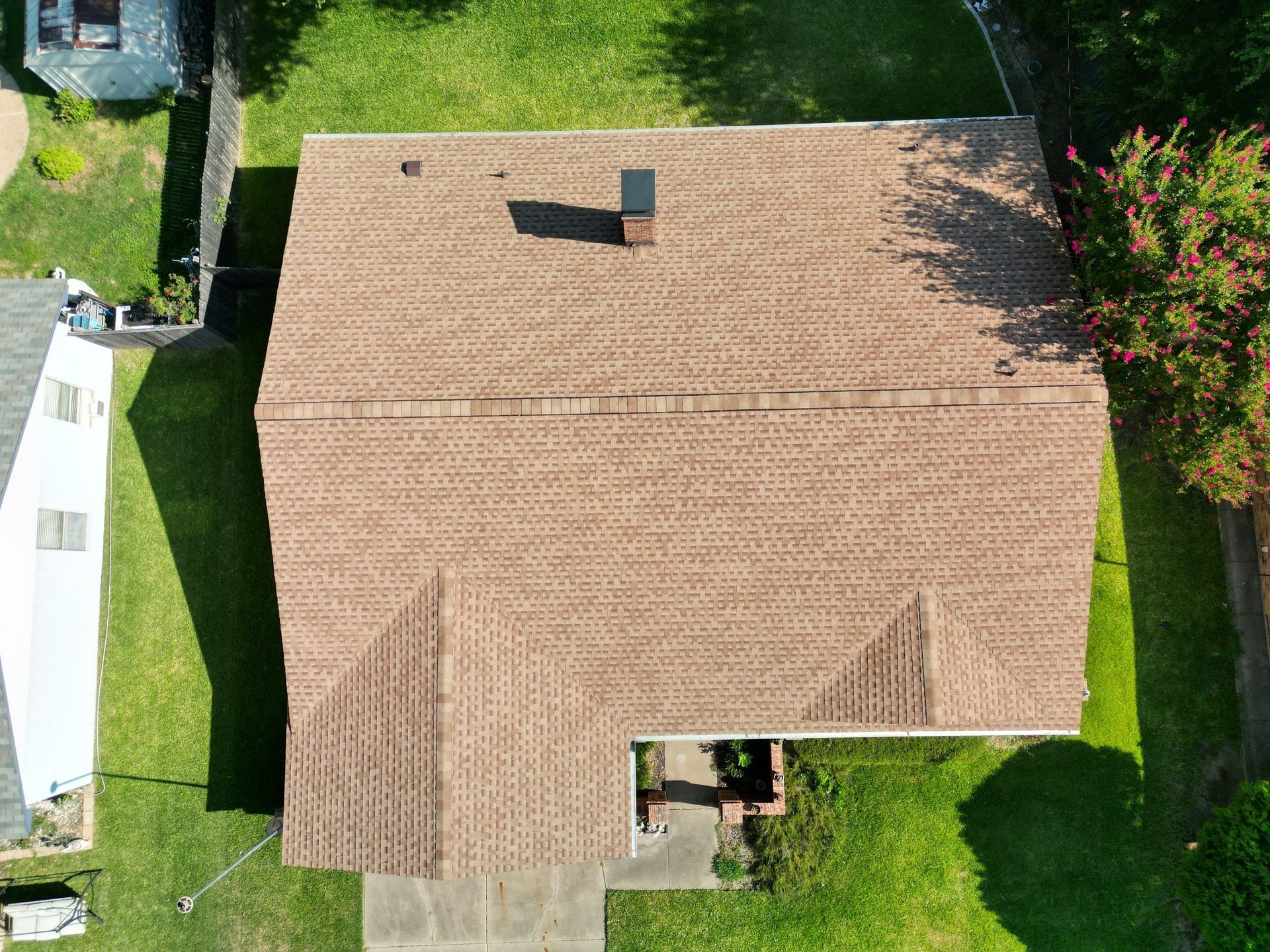 Aerial view of a brown shingled house roof with a chimney, surrounded by green lawn and trees.