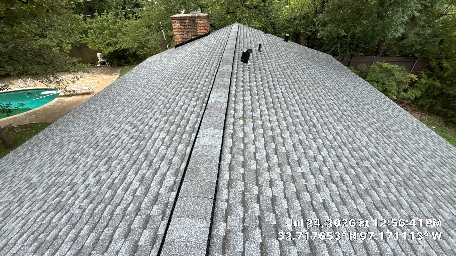 Gray shingled roof with a central ridge seam, seen from above, with trees and a pool in the background