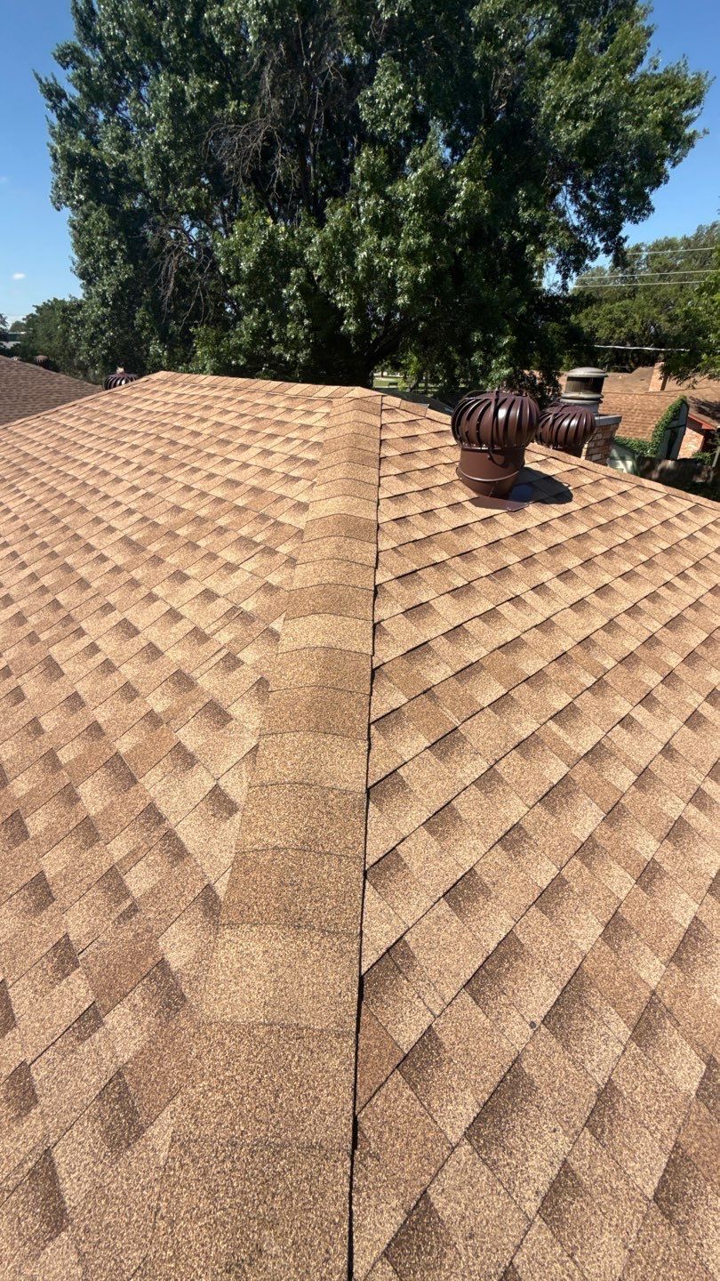 Rooftop with tan shingles and a dark vent pipe, framed by green trees and blue sky.
