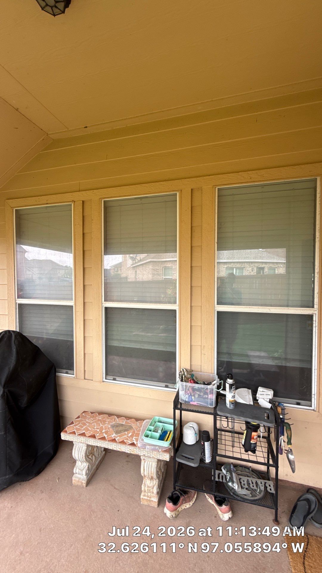 Front porch with three windows, bench, covered grill, and small cart under a yellow overhang.