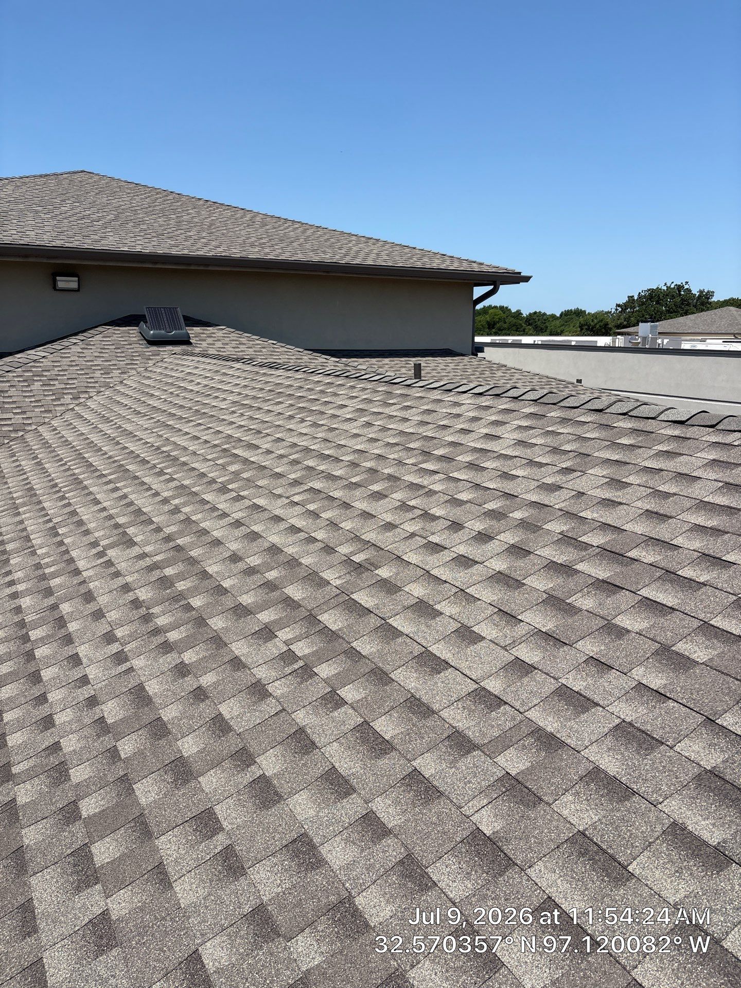 Close-up of a gray shingle roof under a clear blue sky, with a neighboring roof and chimney visible.