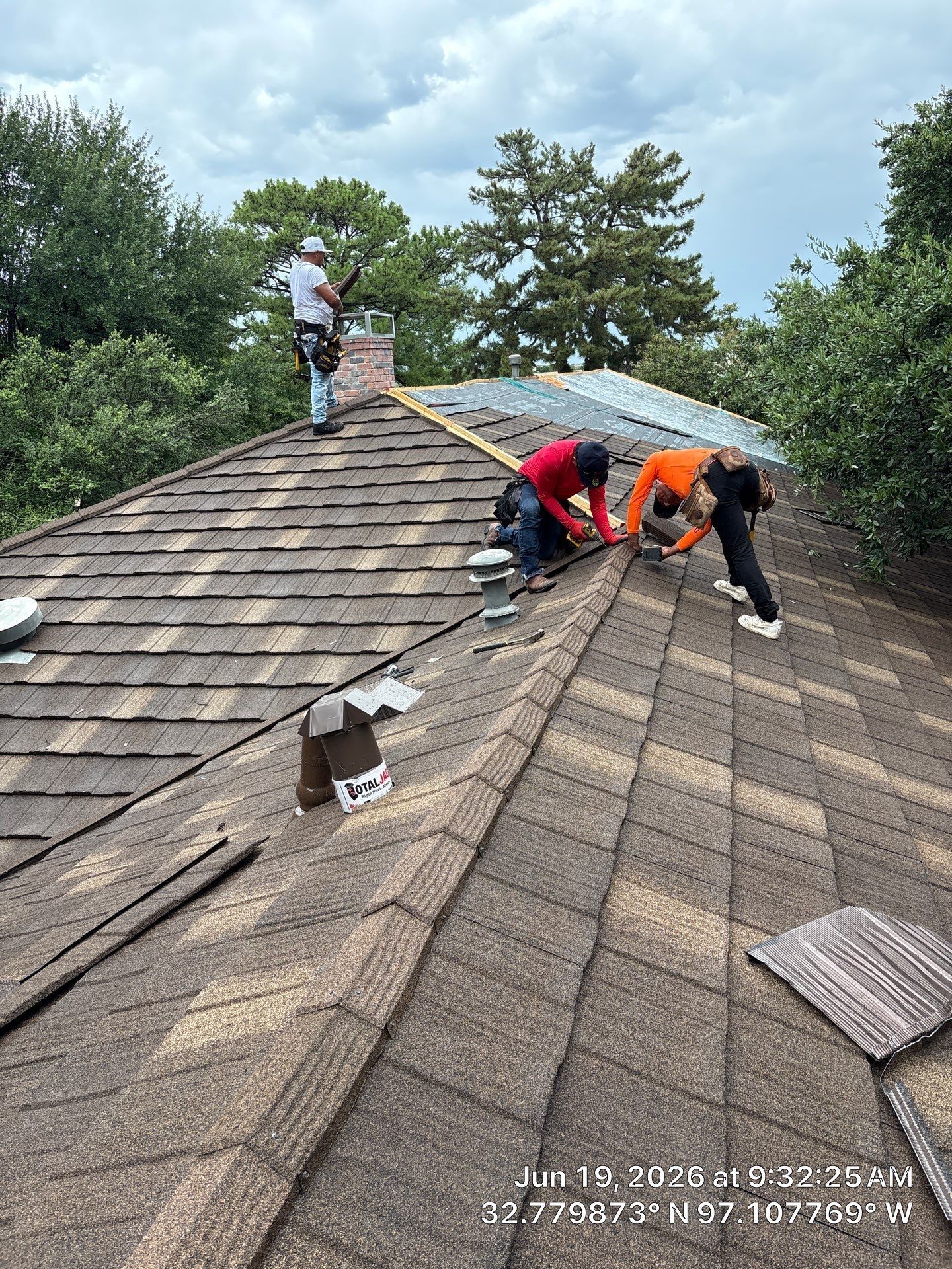 Workers repairing a shingle roof on a house, with one worker on the ridge and two kneeling near the edge.