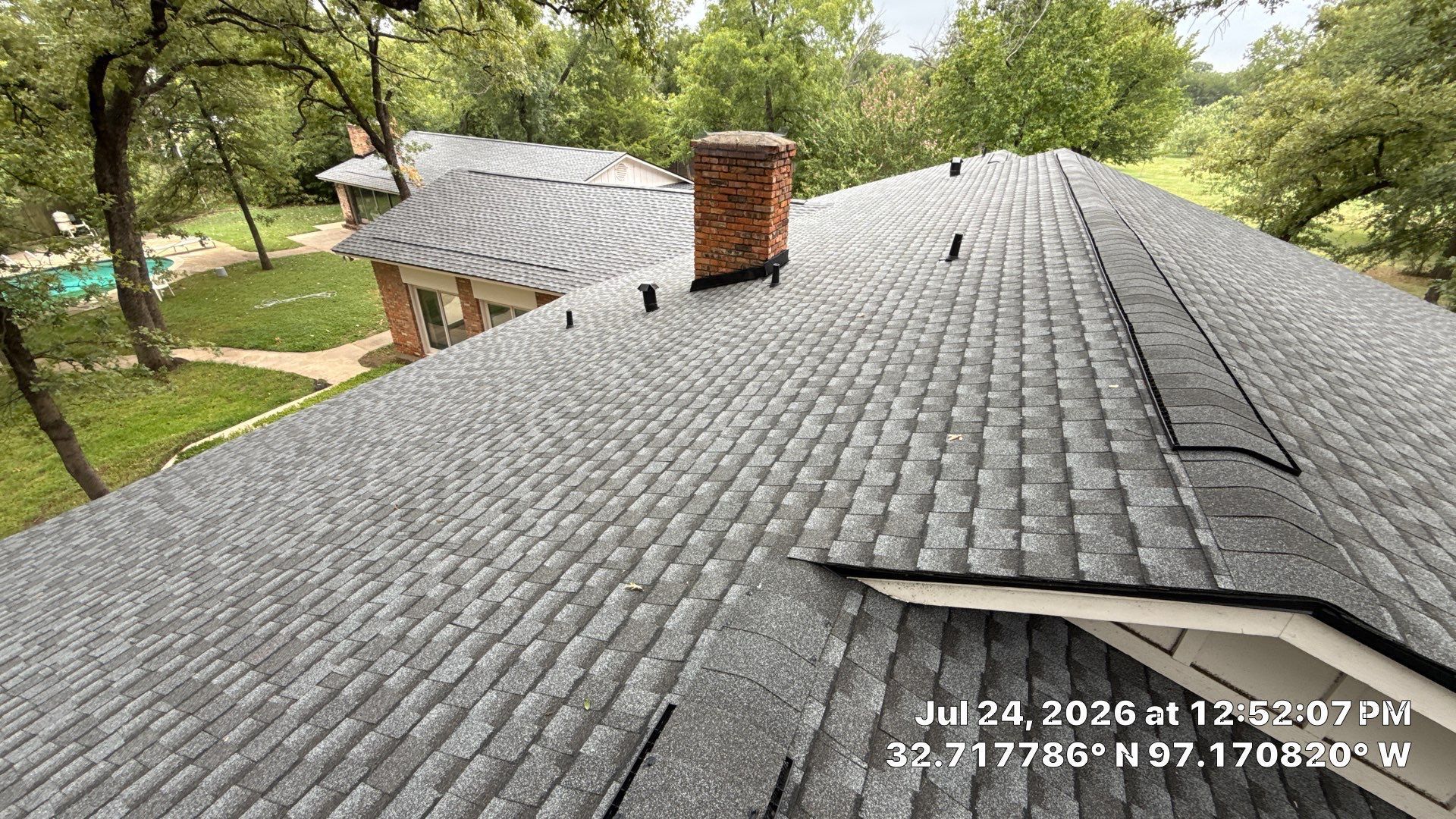 Gray shingle roof with chimney and roof vents, viewed from above, surrounded by trees.