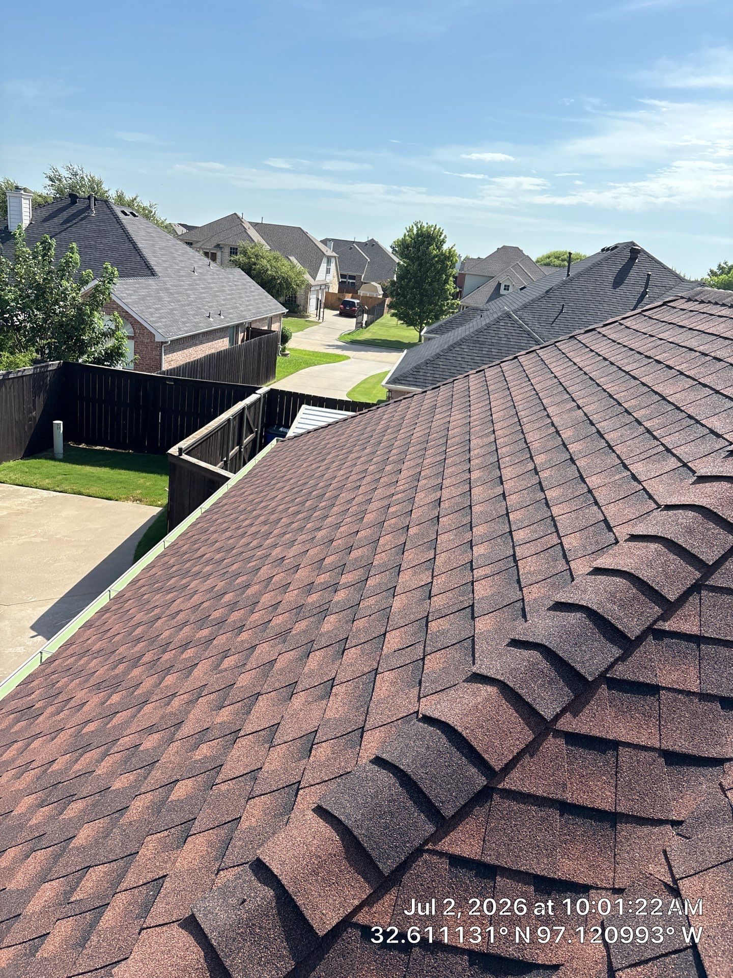 Suburban rooftops with brown shingle roofs and a green lawn under a clear blue sky