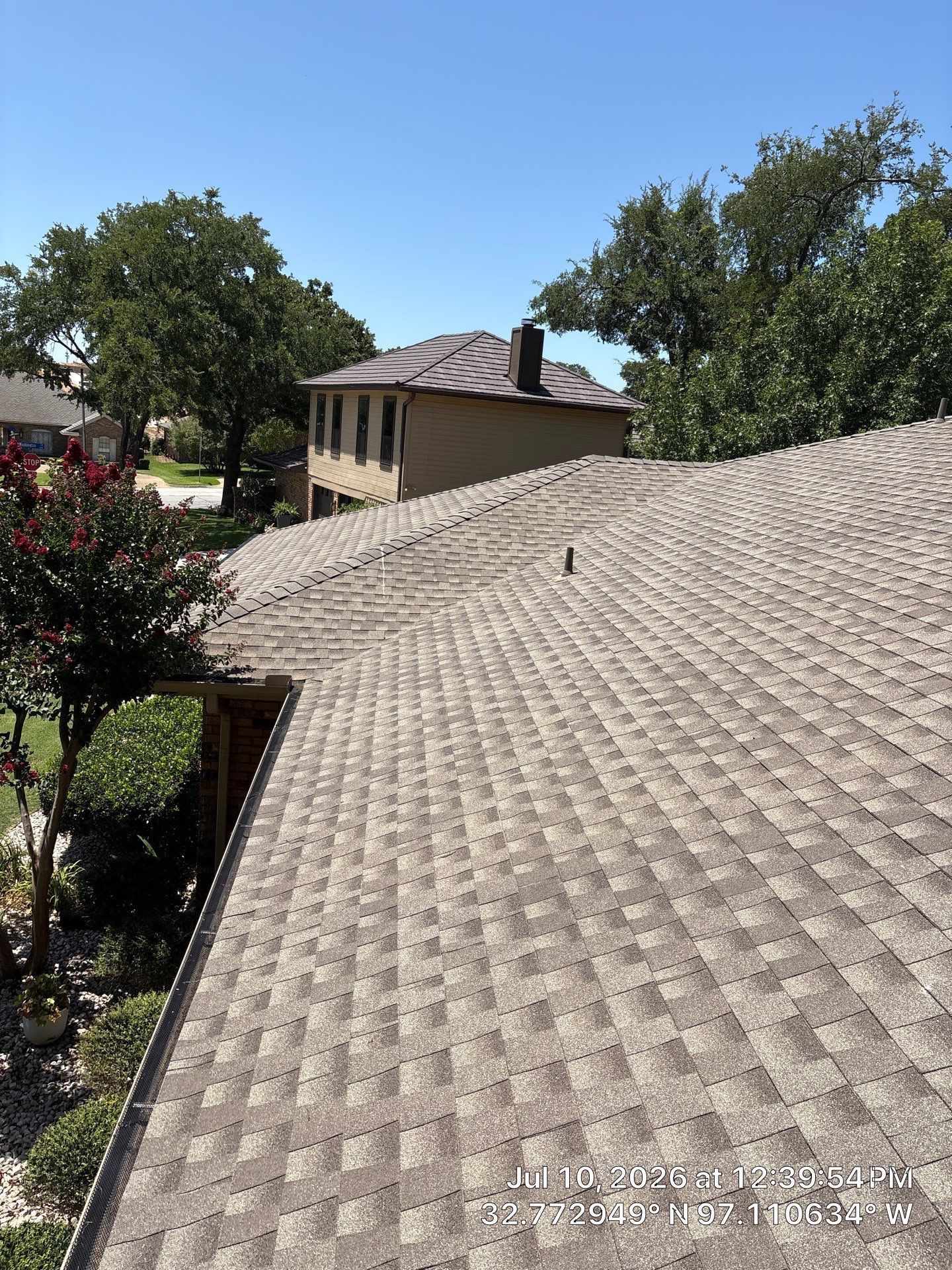 Rooftop shingles in foreground with trees and a small beige house under a bright blue sky