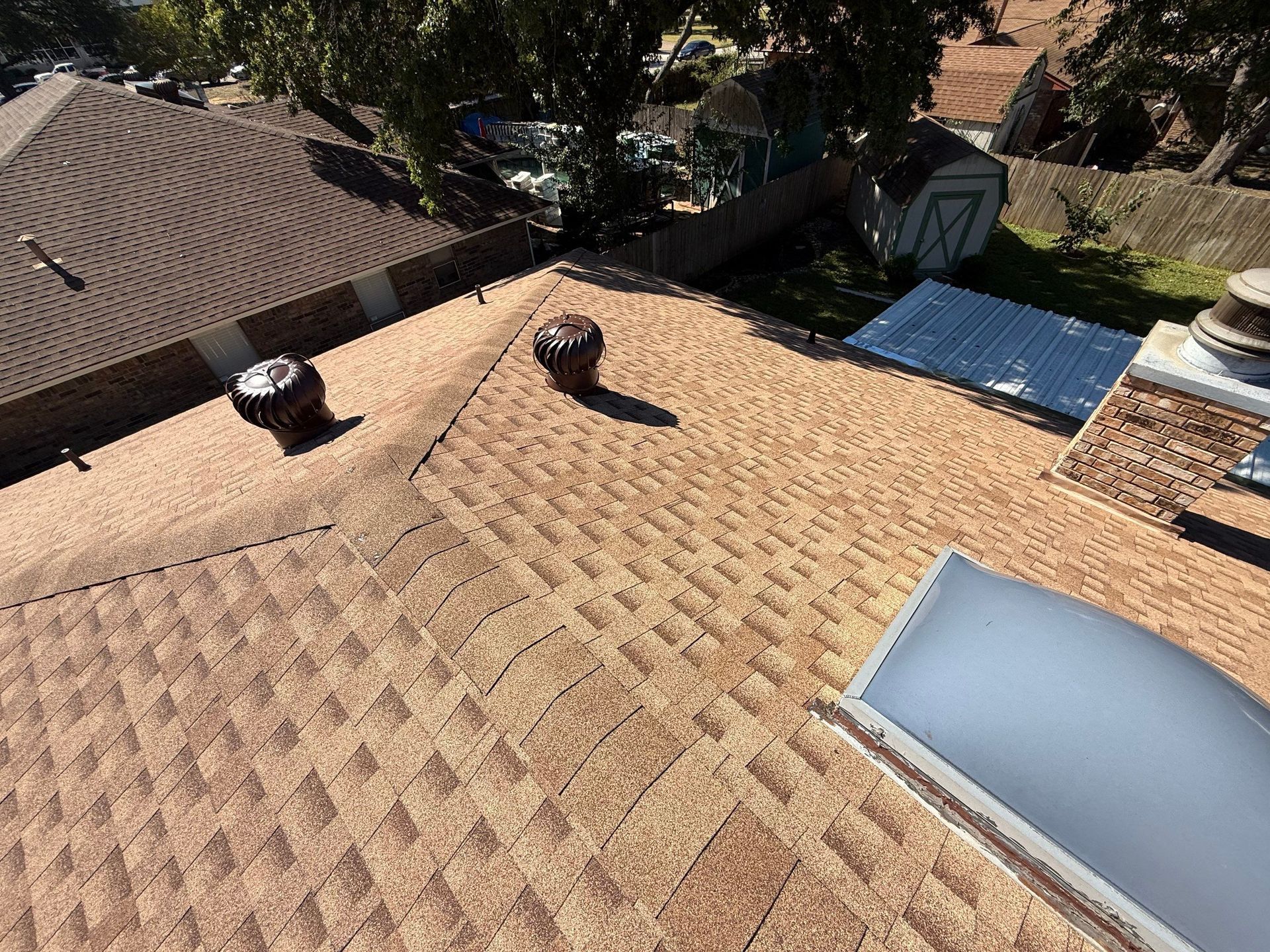 Rooftop with tan shingles, two vent pipes, and a skylight near trees and neighboring houses