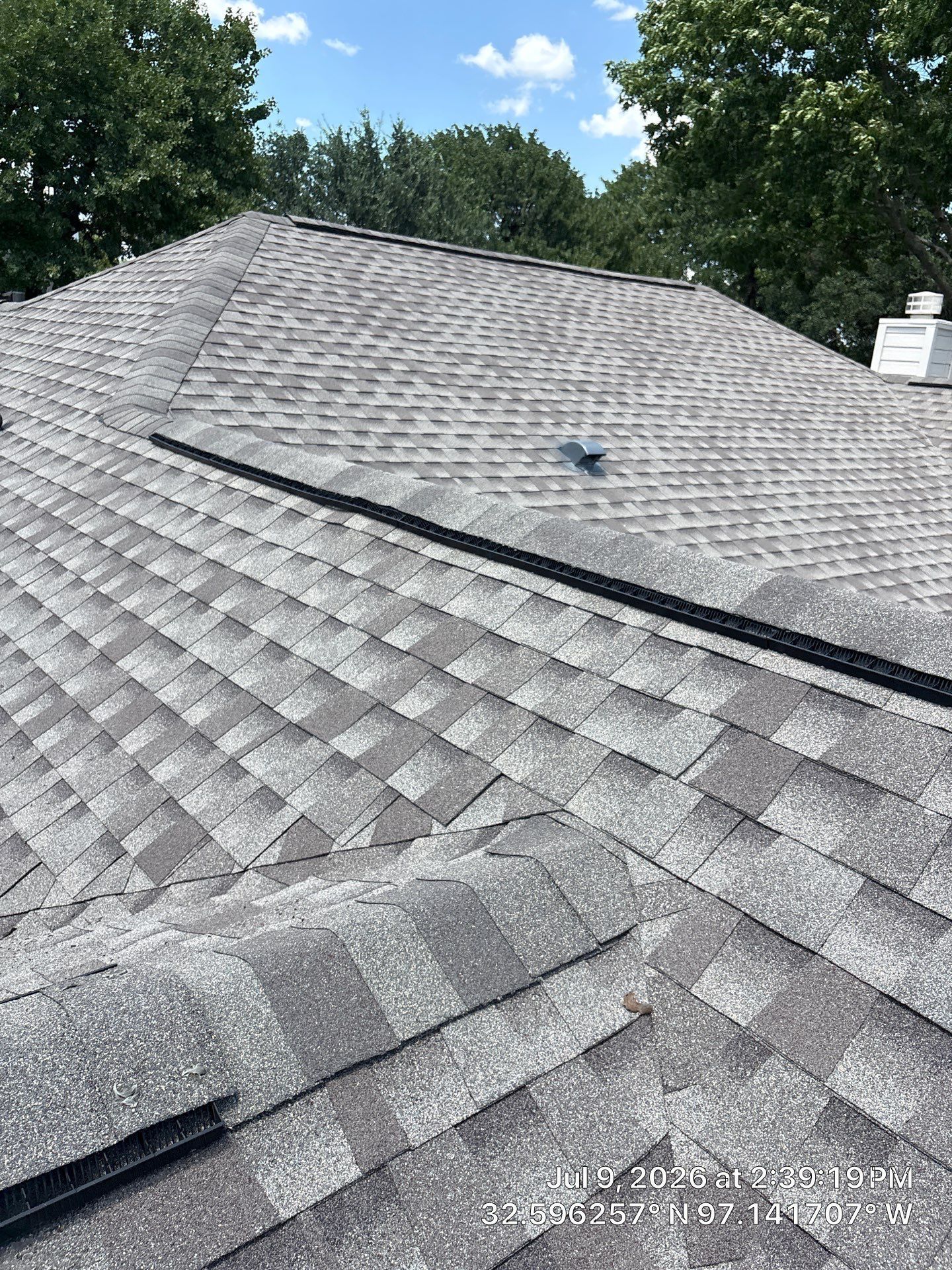 Gray shingled roof with a long dark ridge vent, viewed from above with trees in the background