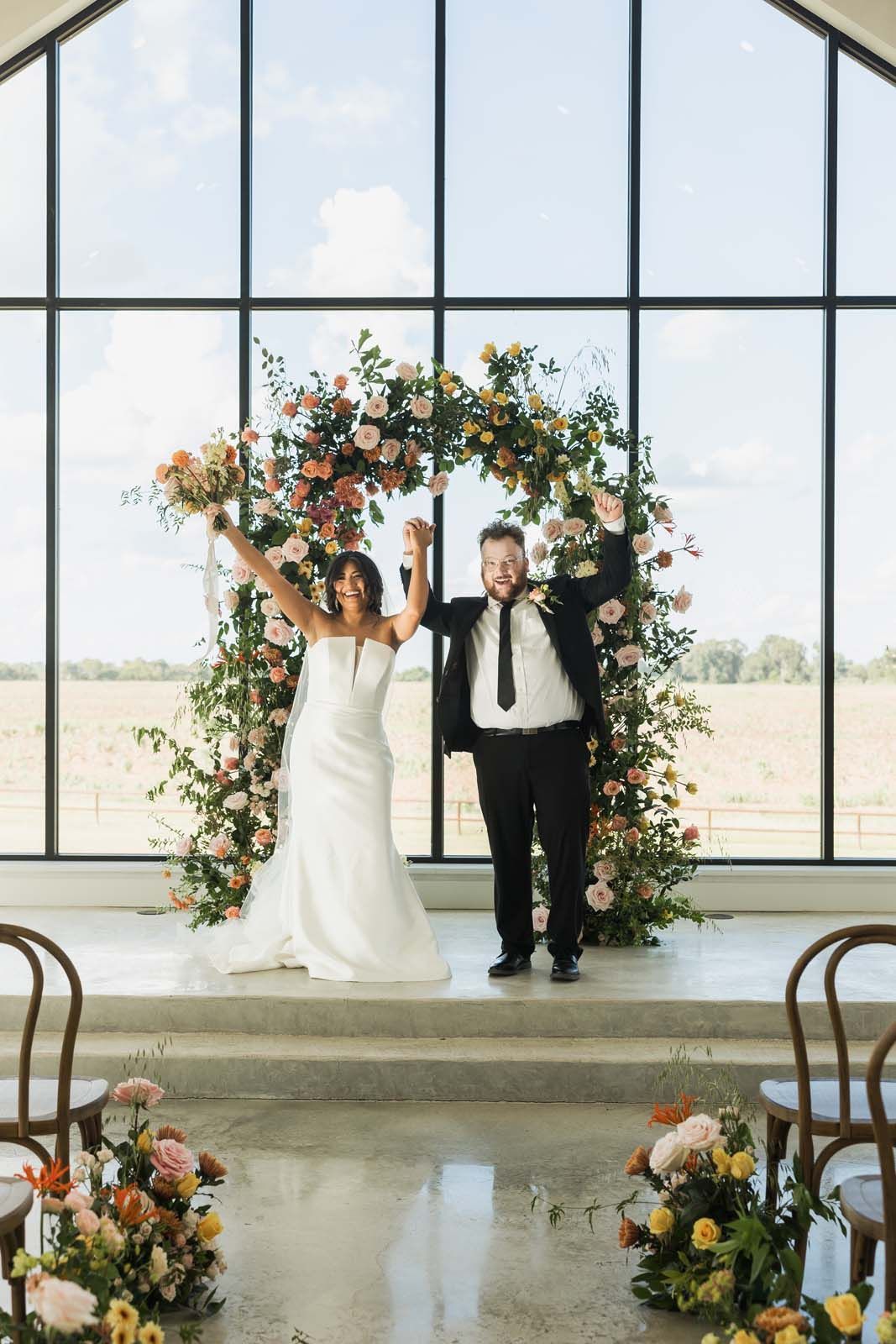 Newlyweds holding hands under a floral arch in a bright, glass-walled venue