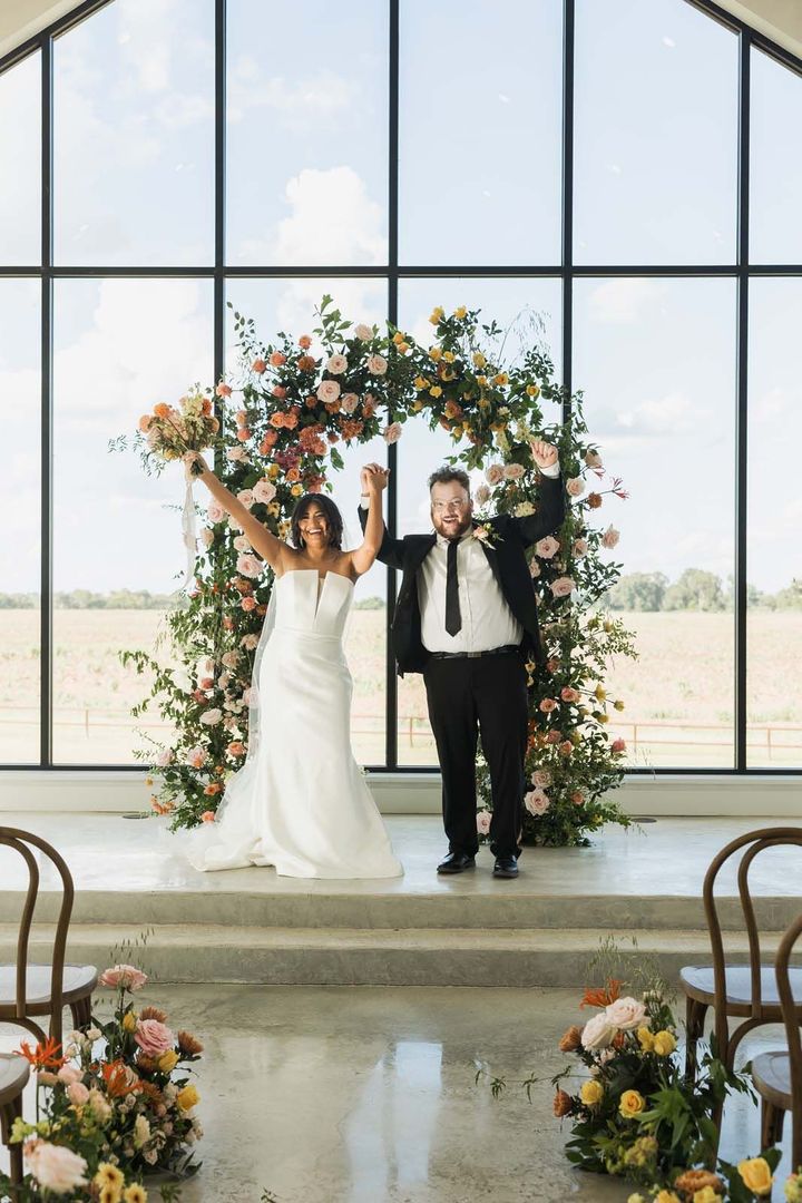 Newlyweds holding hands under a floral arch in a bright, glass-walled venue