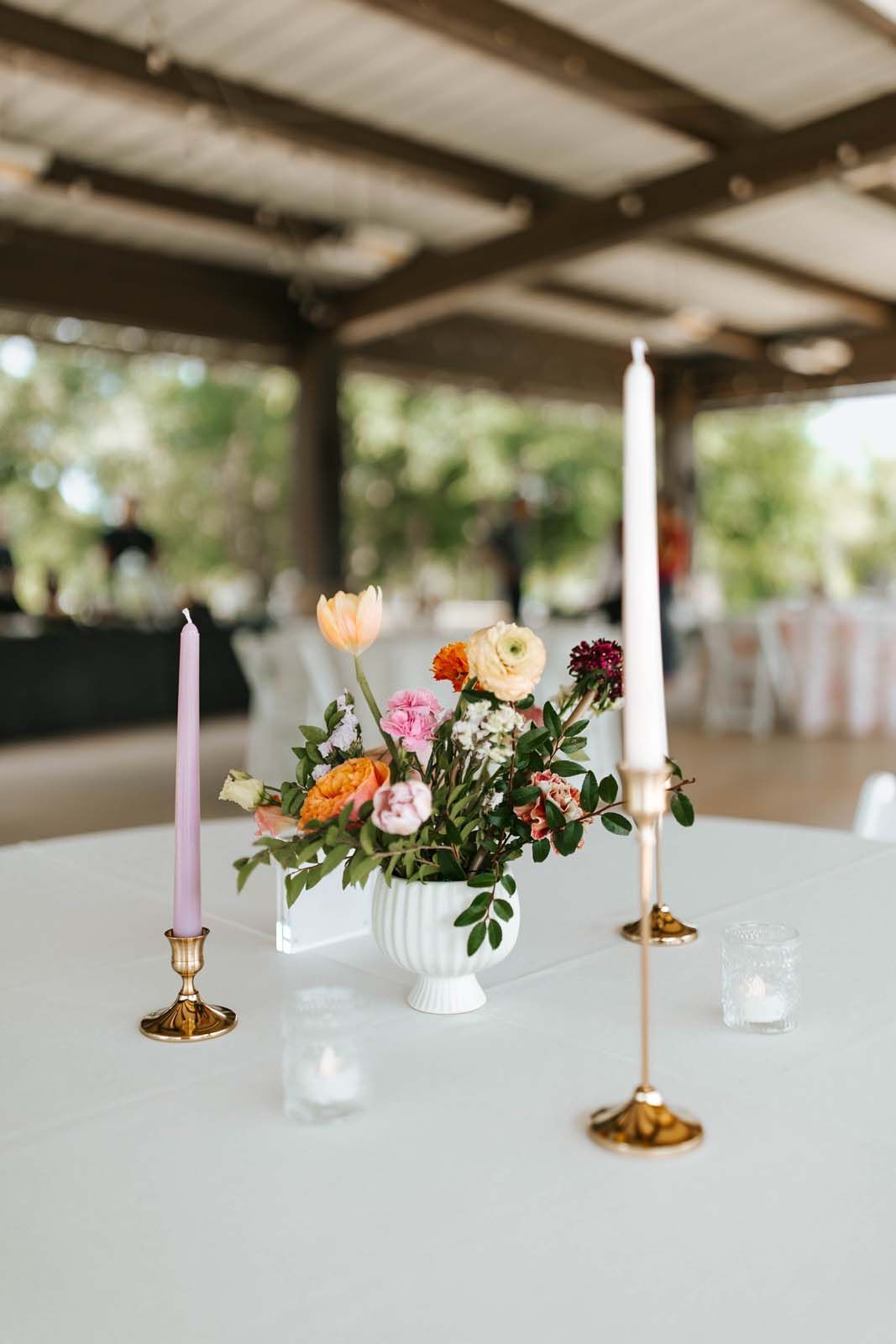 Floral centerpiece with white candles on a round table at an outdoor wedding reception pavilion