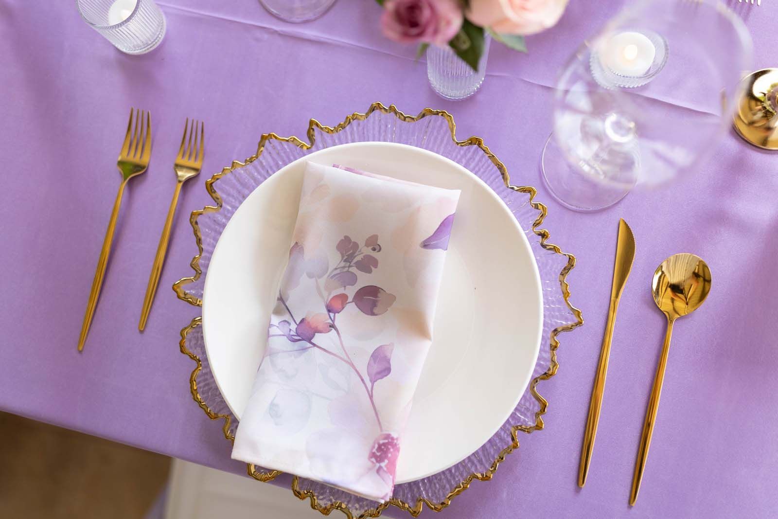 Lavender table setting with gold cutlery, white plates, and floral napkins on a lilac tablecloth