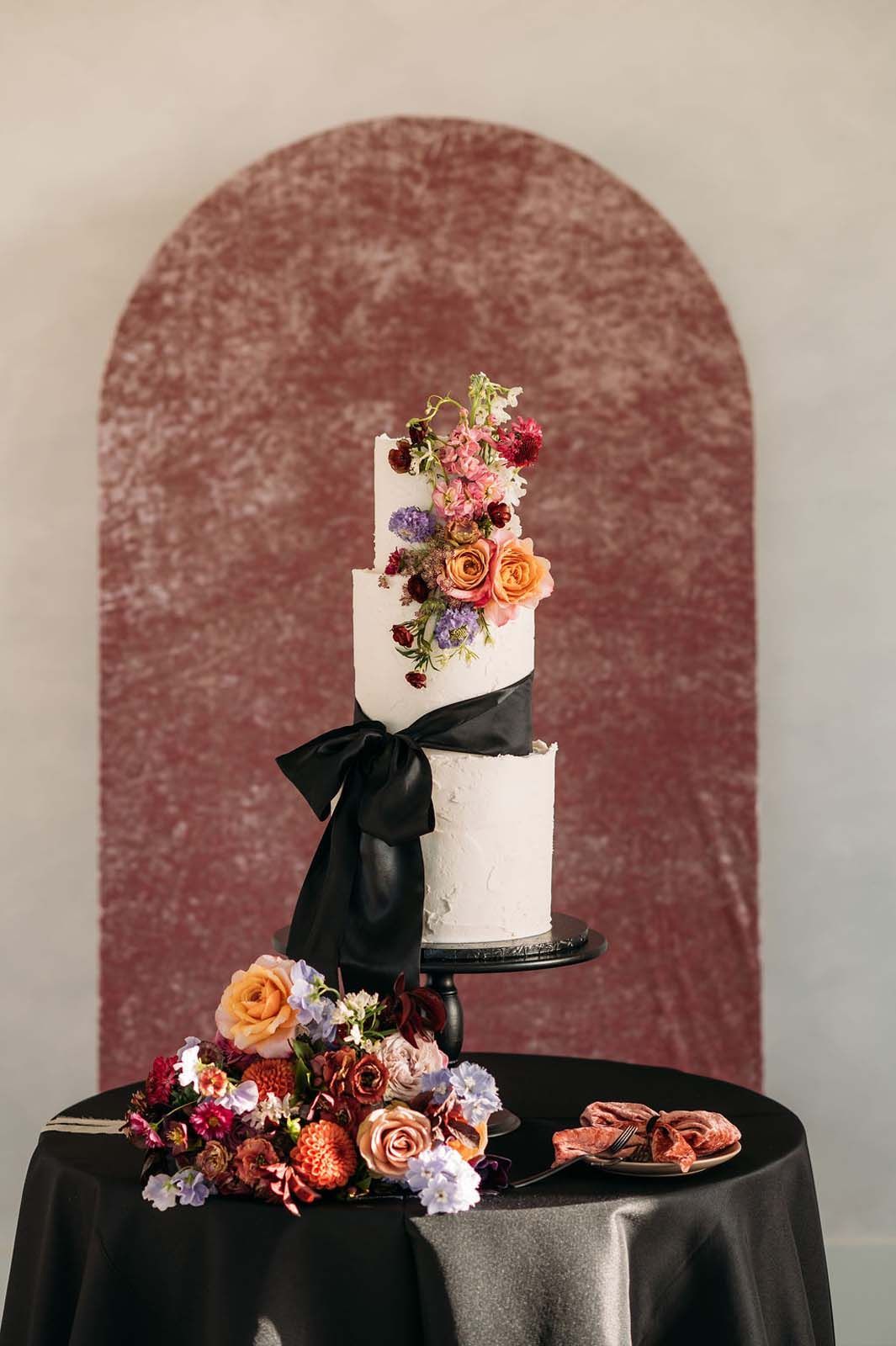 Two-tier white cake with black ribbon and colorful flowers on a black table against a mauve backdrop