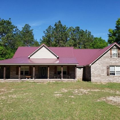 A one-story brick house with a burgundy metal roof, a wrap-around front porch, and trees in the background under a blue sky.