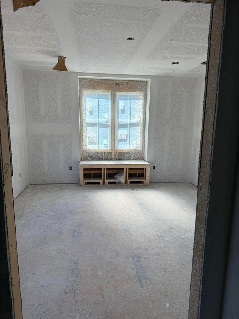 An unfinished room featuring drywall, a central window with a built-in wooden bench, and gray floor covering.