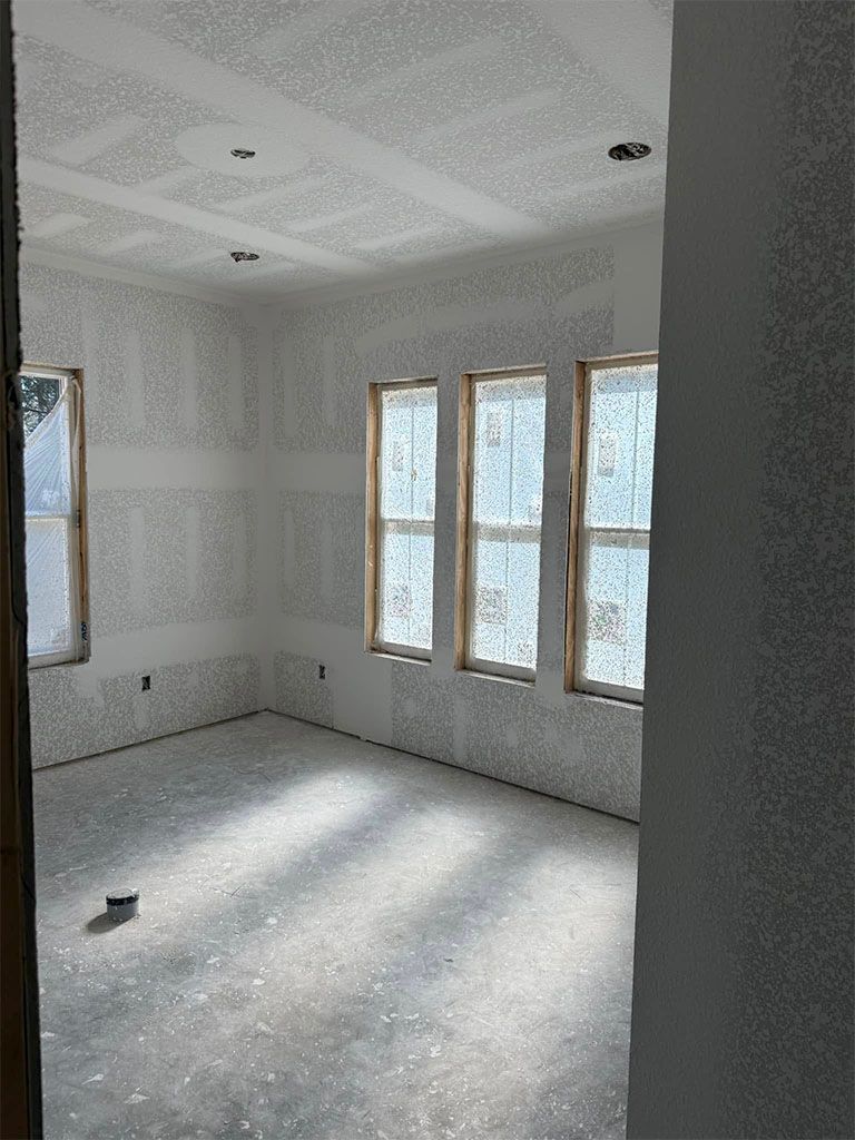 An unfinished room under construction, featuring bare drywall on walls and ceiling, unpainted trim, and large windows.
