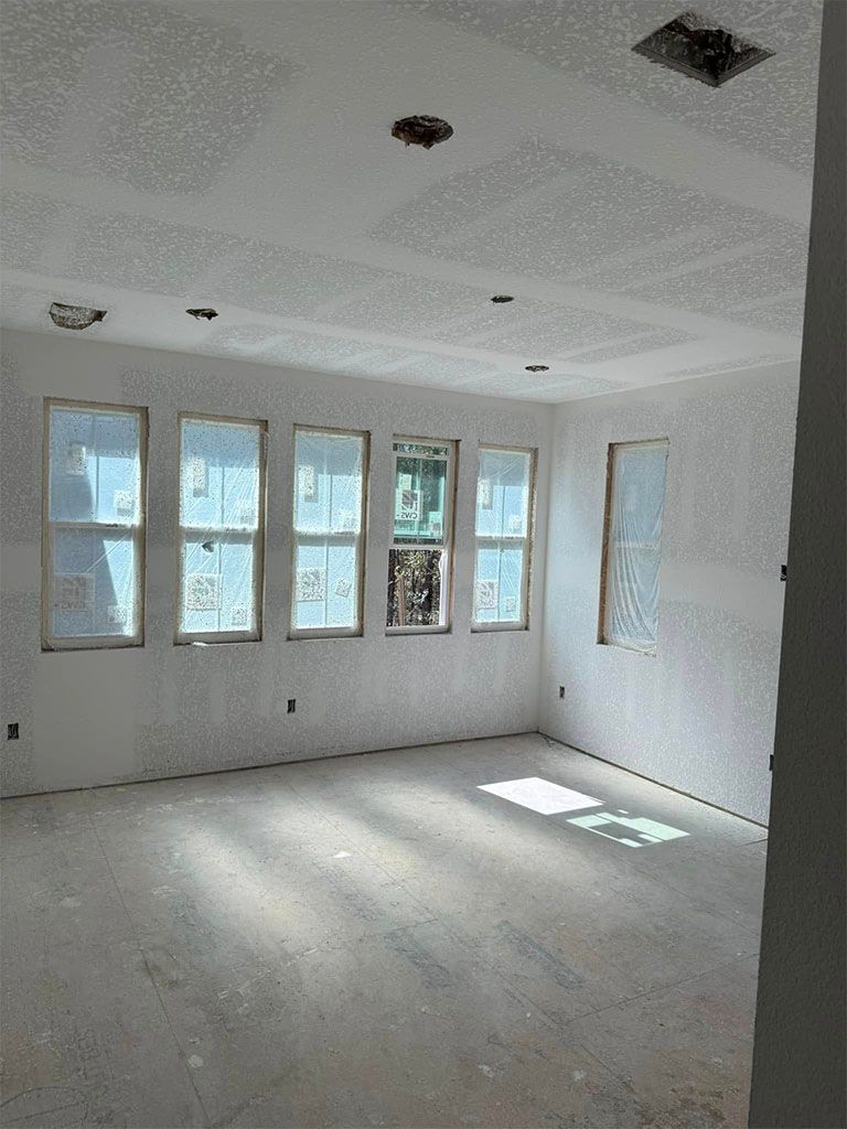 An unfinished, empty room with light grey walls, white ceilings, and a row of large windows letting in sunlight.