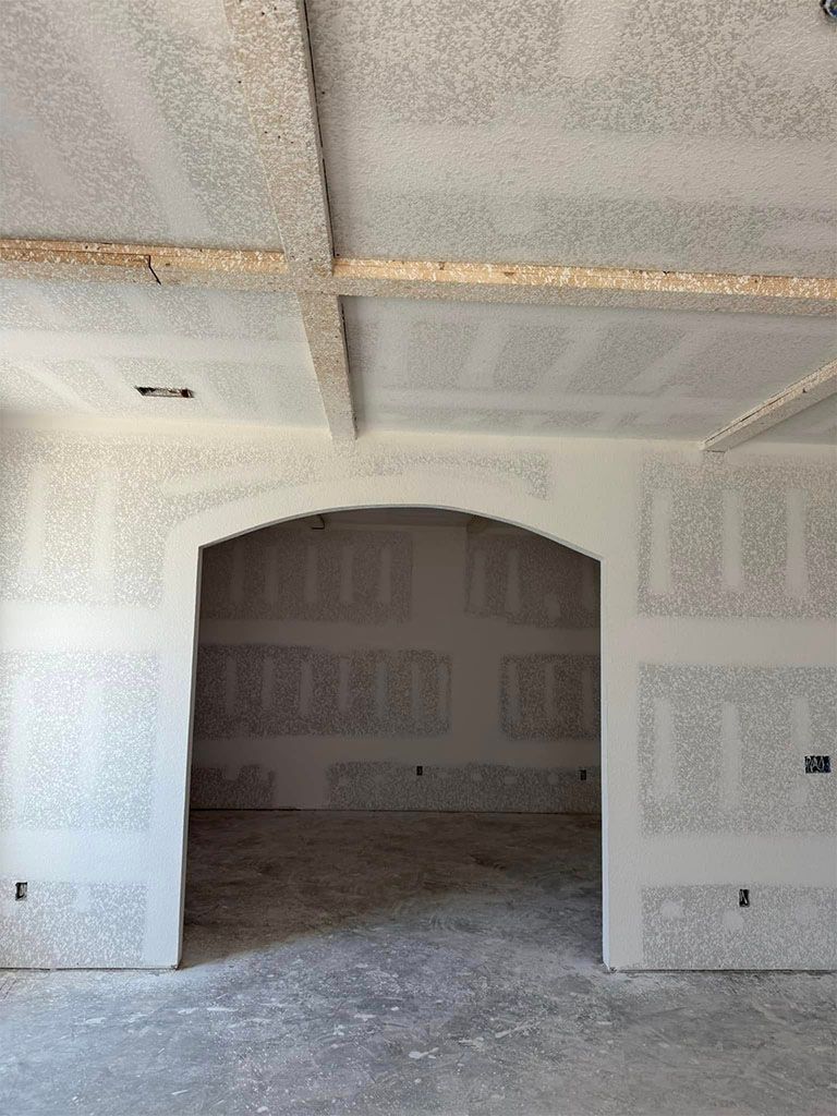 Interior view of a room under construction with unfinished drywall, a coffered ceiling, and a prominent arched entryway.