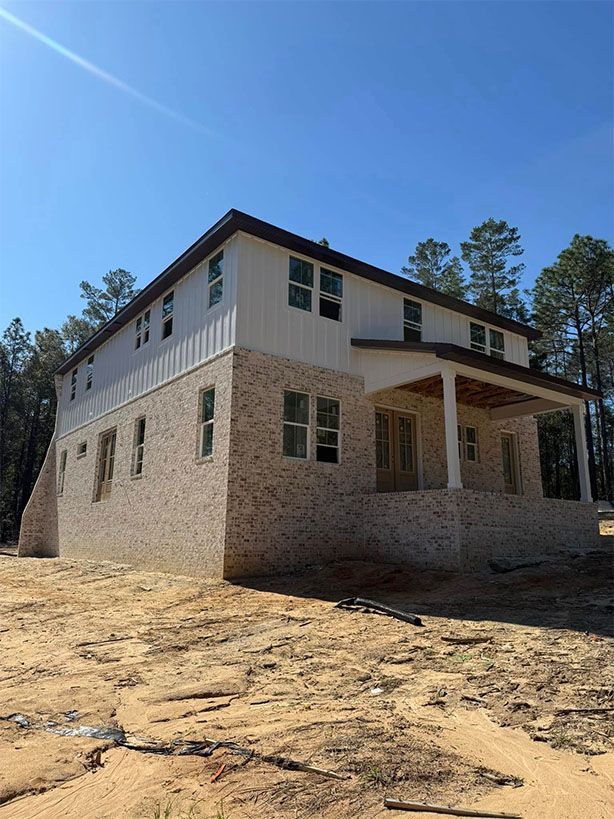 A newly constructed two-story house with a brick bottom floor, white siding, and a covered porch on a dirt lot.
