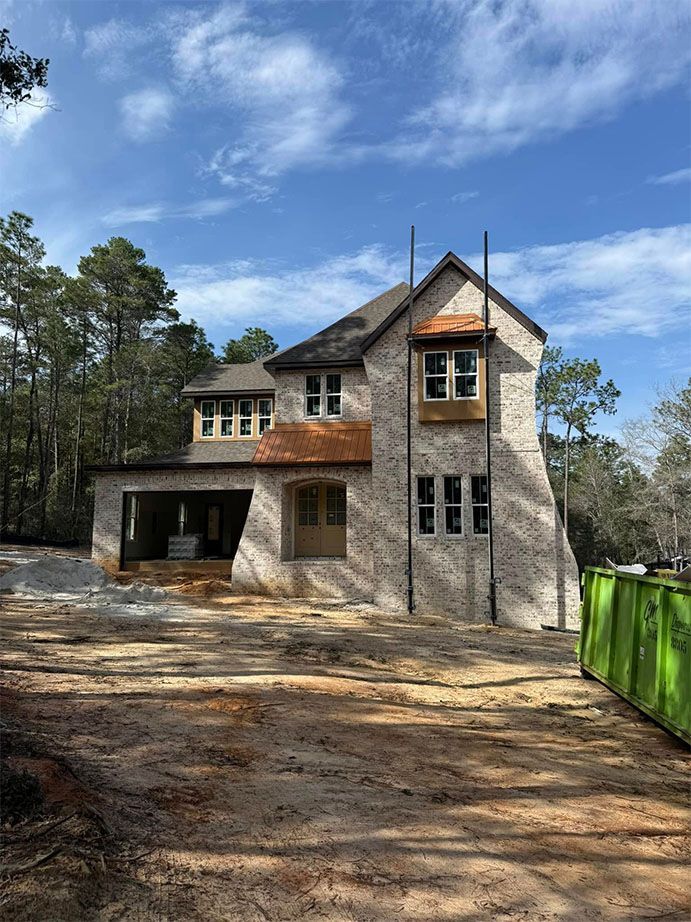 A two-story stone house under construction, featuring a garage, wood-trimmed windows, and a green dumpster in the yard.