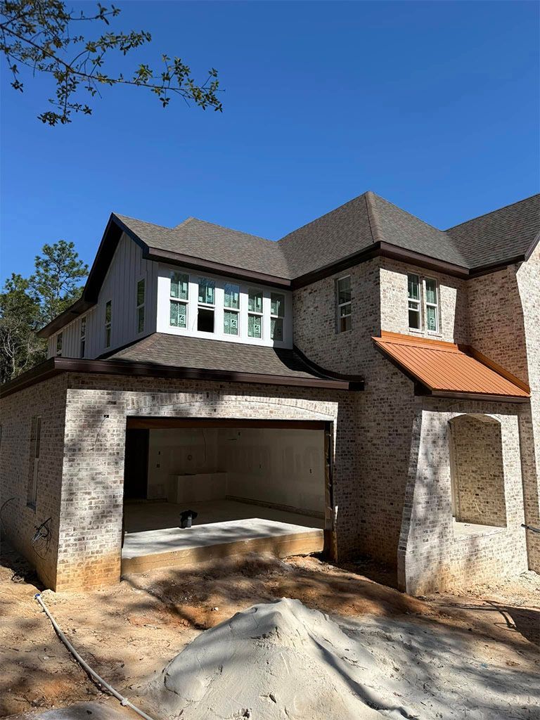 A newly constructed, two-story house with white brick exterior, dark shingles, and an open garage bay under a clear sky.