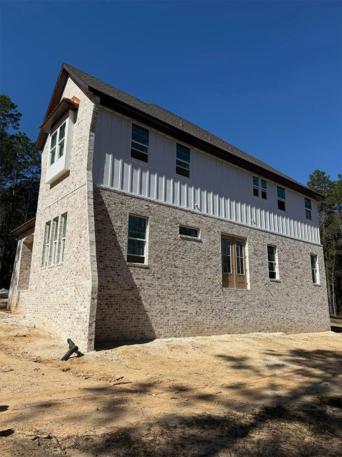 Two-story house under construction with light-colored brick siding, white vertical siding, and a dark shingled roof.