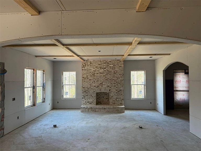 A construction-phase room featuring an exposed brick fireplace, arched doorway, and unfinished drywall and ceiling beams.