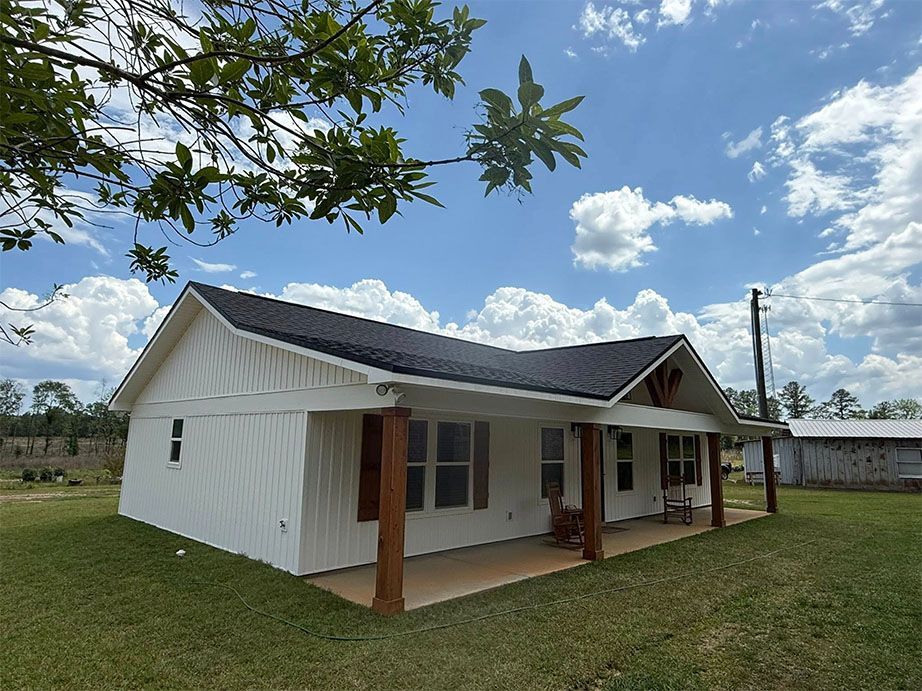 A white farmhouse with dark wood accents and a porch, situated in a grassy field under a bright, cloudy sky.