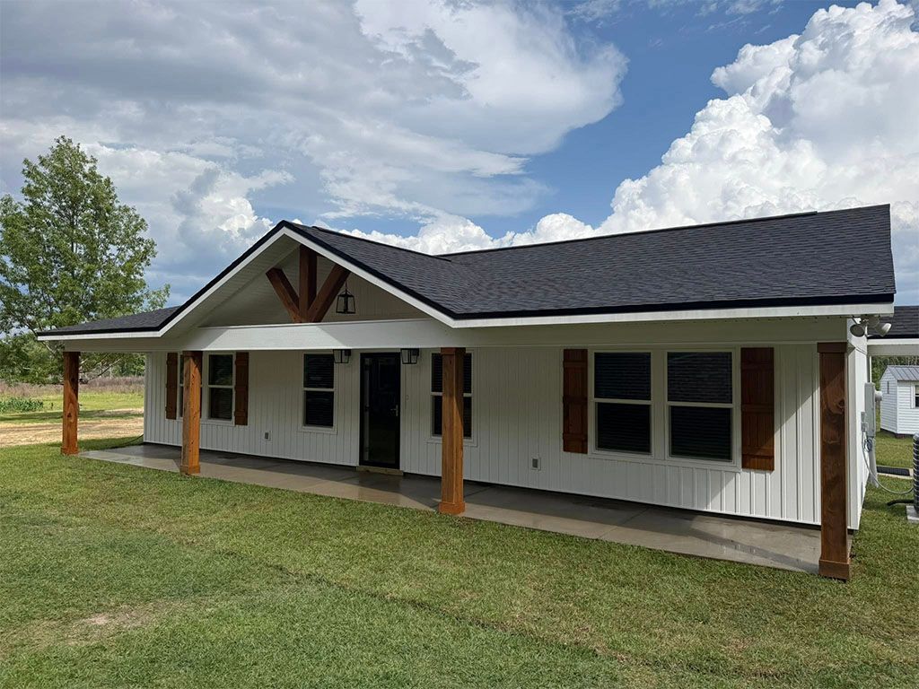 Single-story white house with board-and-batten siding, a dark roof, wooden porch posts, and wood-toned window shutters.