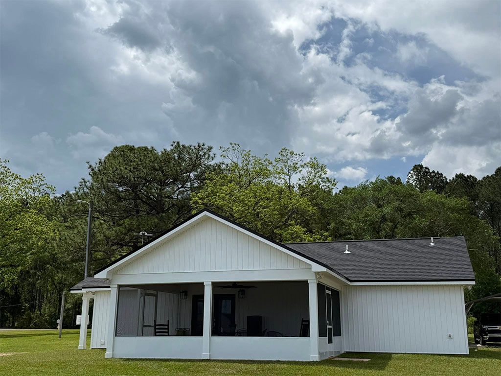 White farmhouse-style home with a screened-in porch, dark shingled roof, and green trees under a cloudy sky.