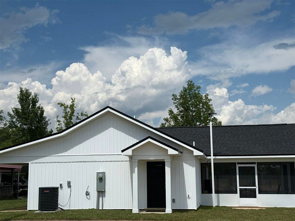 White house with a black roof, front porch, and a screened-in sunroom under a bright blue sky with puffy white clouds.