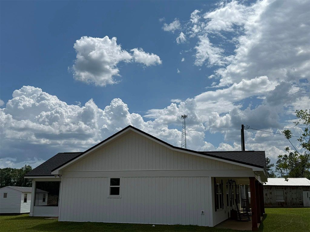 A white house with a dark roof stands under a blue sky filled with large, bright white cumulus clouds.