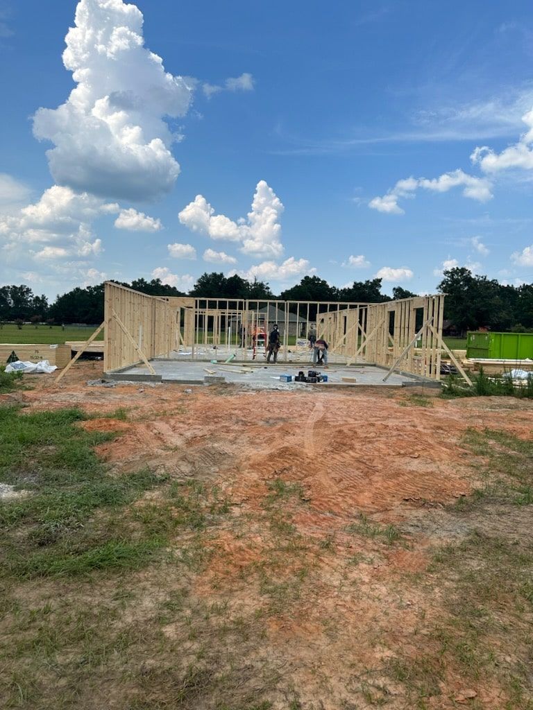 A wooden wall frame under construction on a concrete foundation in a sunny, open field under a blue, cloudy sky.