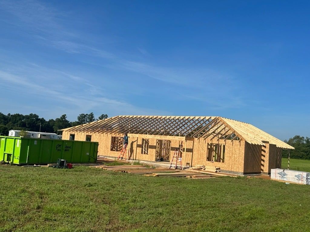 A wooden house frame under construction in a grassy field with a large green dumpster to the side under a clear blue sky.