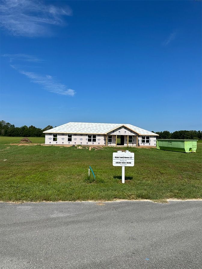 A single-story house under construction with white exterior sheathing, surrounded by a grassy field under a blue sky.
