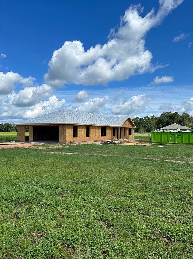 A new wood-framed house under construction stands in a grassy field under a bright blue sky with scattered clouds.