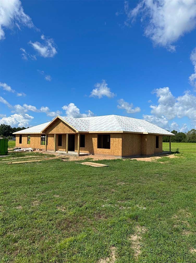 A newly framed, single-story house with a white roof under construction on a sunny day in an open, grassy field.