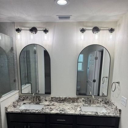 A bathroom vanity with two arched mirrors, dark cabinets, granite countertops, and two three-light fixtures above.