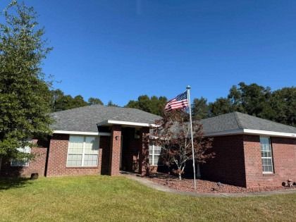 A one-story brick house with a gray shingled roof, a front porch, and an American flag on a pole in the yard.