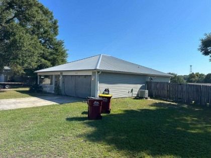 A detached, light-gray garage with a metal roof stands on a grassy lawn next to a wooden fence on a sunny day.
